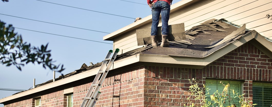 Contractor works on a roofing project, standing on the roof with a ladder along the side of the house.